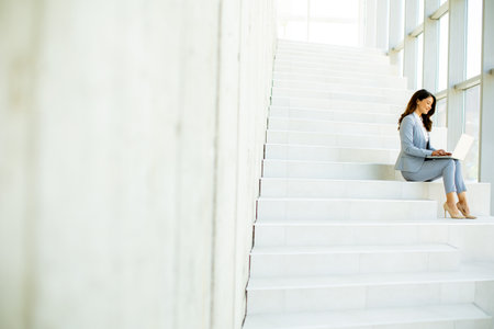 Pretty young business woman sitting on the stairs on office hallway and working on laptopの写真素材