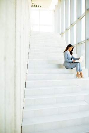 Pretty young business woman sitting on the stairs on office hallway and working on laptopの写真素材