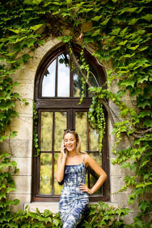 Pretty young woman using mobile phone by old house with ivyの写真素材