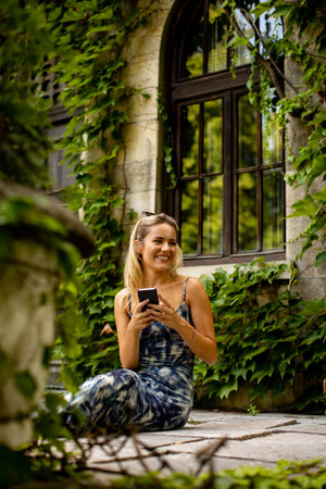Pretty young woman using mobile phone by old house with ivyの写真素材