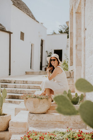 A young woman in a white dress and  hat relaxing on stairs on a sunny day during tourist visit in Alberobello, Italyの写真素材