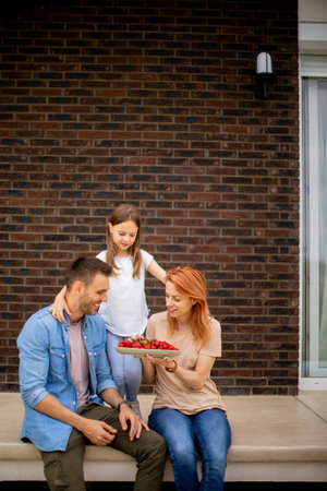 Family with a mother, father and daughter sitting outside on a steps of a front porch of a brick house and eating strawberriesの写真素材