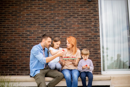 Family with a mother, father, son and daughter sitting outside on a steps of a front porch of a brick house and eating strawberriesの写真素材