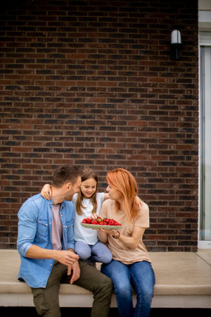 Family with a mother, father and daughter sitting outside on a steps of a front porch of a brick house and eating strawberriesの写真素材