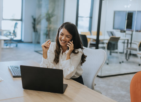 Pretty young business woman using mobile phone while working on laptop in the modern officeの写真素材