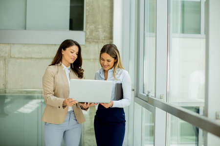 Two pretty young business women with laptop in the office hallwayの写真素材