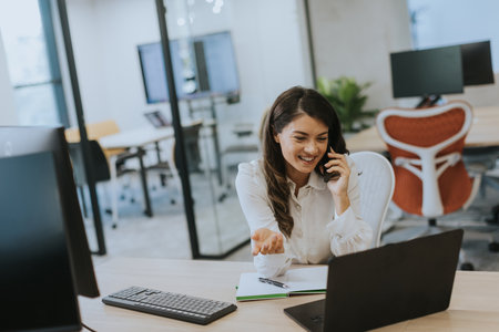Pretty young business woman using mobile phone while working on laptop in the modern officeの写真素材
