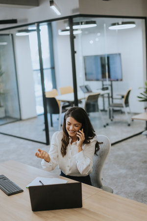 Pretty young business woman using mobile phone while working on laptop in the modern officeの写真素材