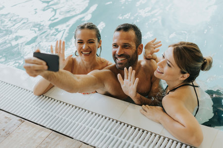 Handsome young people relaxing in the indoor swimming pool and taking selfie with mobile phoneの写真素材