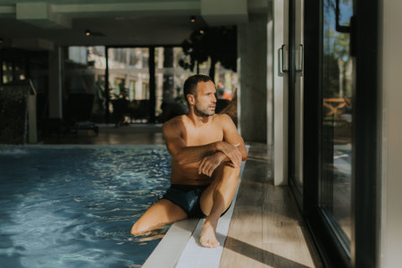 Handsome young man relaxing on the edge of a indoor swimming poolの写真素材