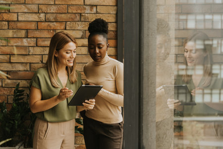 Two pretty young business women with digital tablet standing by the brick wall in the industrial style officeの写真素材
