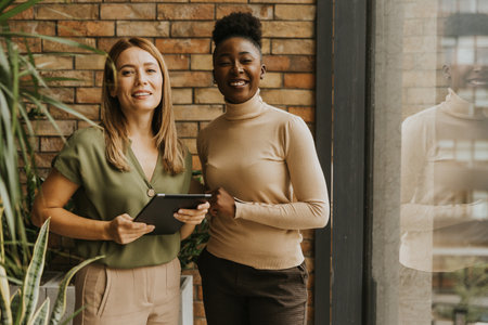 Two pretty young business women with digital tablet standing by the brick wall in the industrial style officeの写真素材