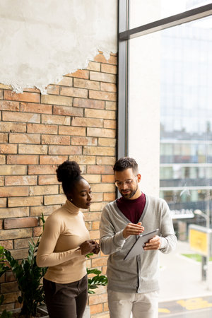 Handsome young multiethnic business couple with digital tablet standing and discussing in the modern officeの写真素材