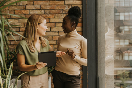 Two pretty young business women with digital tablet standing by the brick wall in the industrial style officeの写真素材
