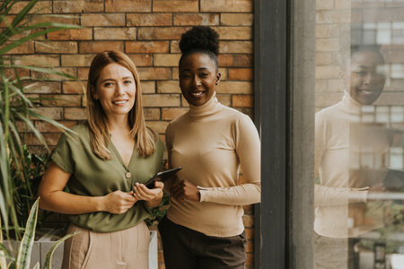 Two pretty young business women with digital tablet standing by the brick wall in the industrial style officeの写真素材