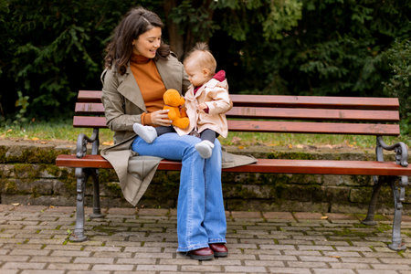 Young woman sitting on a bench with a cute baby girl in the autumn parkの写真素材