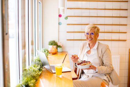 Good looking senior woman working on a laptop, drinking orange juice and having a healthy breakfast in cafeteriaの写真素材