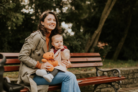Young woman sitting on a bench with a cute baby girl in the autumn parkの写真素材