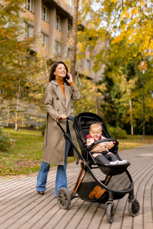 Young woman with a cute baby girl in baby stroller using mobile phone at the autumn parkの写真素材