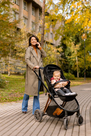 Young woman with a cute baby girl in baby stroller using mobile phone at the autumn parkの写真素材