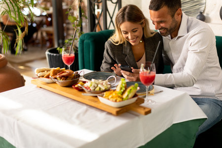 Young couple using mobile phone while having a lunch and drinking fresh squeezed juice in the restaurantの写真素材