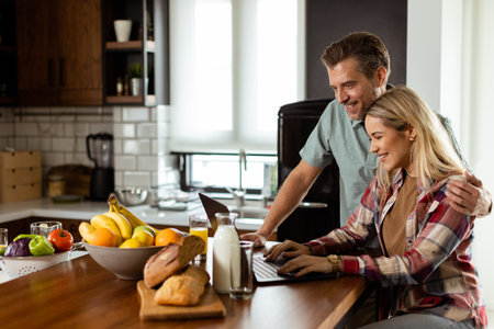 Smiling couple shares a moment of togetherness as they sit by the kitchen counter, surrounded by a spread of fresh fruits and pastries, bathed in the warm morning light while browsing on a laptopの写真素材