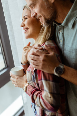 In a softly lit room, a man lovingly embraces a woman from behind as they both gaze out the window, sharing a quiet, intimate moment togetherの写真素材