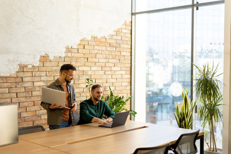 Two smiling professionals engage in a collaborative work session at a wooden table, their camaraderie evident in a contemporary office setting with an exposed brick wall backdropの写真素材
