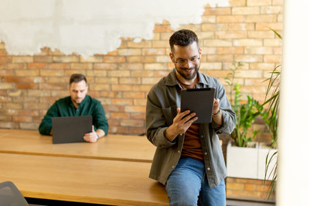 In a contemporary office setting, two focused colleagues engage in their work with tablet and laptop, collaborating and sharing ideas in a bright and casual environment.の写真素材