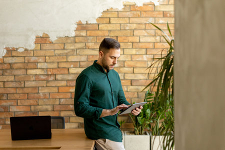 Cheerful man stands confidently holding a digital tablet in a contemporary office space with an exposed brick wall, symbolizing a blend of modern tech and rustic charmの写真素材