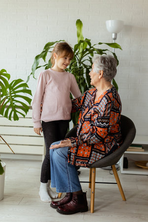 In a softly-lit room, a young girl stands beside a potted plant, sharing a moment of connection and conversation with her elderly grandmother, the joy and warmth between them palpableの写真素材