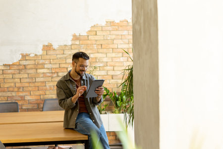 Cheerful man stands confidently holding a digital tablet in a contemporary office space with an exposed brick wall, symbolizing a blend of modern tech and rustic charmの写真素材
