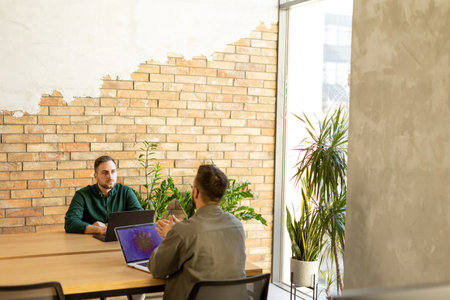 Two smiling professionals engage in a collaborative work session at a wooden table, their camaraderie evident in a contemporary office setting with an exposed brick wall backdropの写真素材
