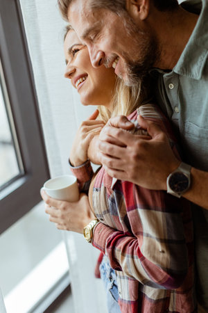 In a softly lit room, a man lovingly embraces a woman from behind as they both gaze out the window, sharing a quiet, intimate moment togetherの写真素材