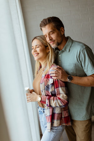 In a softly lit room, a man lovingly embraces a woman from behind as they both gaze out the window, sharing a quiet, intimate moment togetherの写真素材