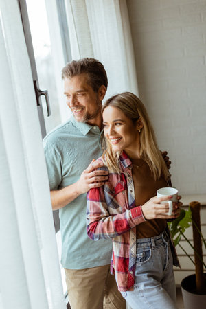 In a softly lit room, a man lovingly embraces a woman from behind as they both gaze out the window, sharing a quiet, intimate moment togetherの写真素材