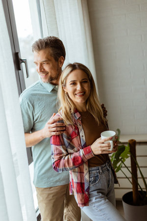In a softly lit room, a man lovingly embraces a woman from behind as they both gaze out the window, sharing a quiet, intimate moment togetherの写真素材