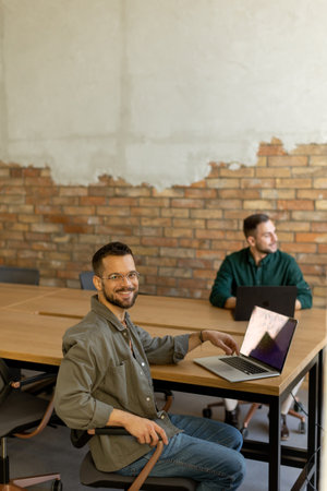 Two smiling professionals engage in a collaborative work session at a wooden table, their camaraderie evident in a contemporary office setting with an exposed brick wall backdropの写真素材