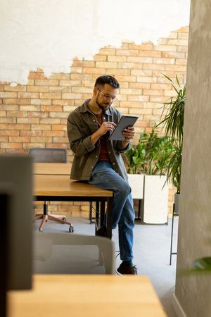 Cheerful man stands confidently holding a digital tablet in a contemporary office space with an exposed brick wall, symbolizing a blend of modern tech and rustic charmの写真素材
