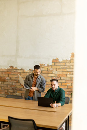 Two smiling professionals engage in a collaborative work session at a wooden table, their camaraderie evident in a contemporary office setting with an exposed brick wall backdropの写真素材