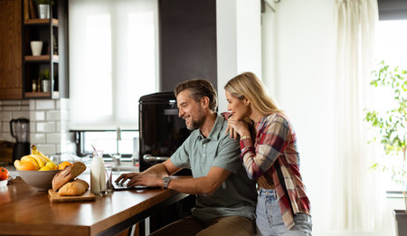 Smiling couple shares a moment of togetherness as they sit by the kitchen counter, surrounded by a spread of fresh fruits and pastries, bathed in the warm morning light while browsing on a laptopの写真素材