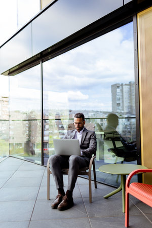 A focused man in a suit uses his laptop on a sunny balcony overlooking the cityscape.の写真素材