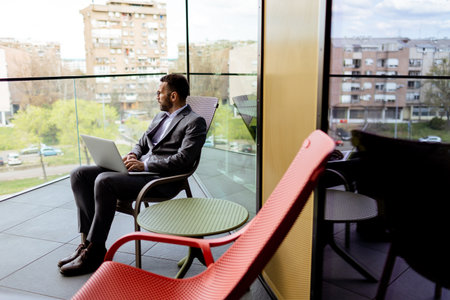 A focused man in a suit uses his laptop on a sunny balcony overlooking the cityscape.の写真素材