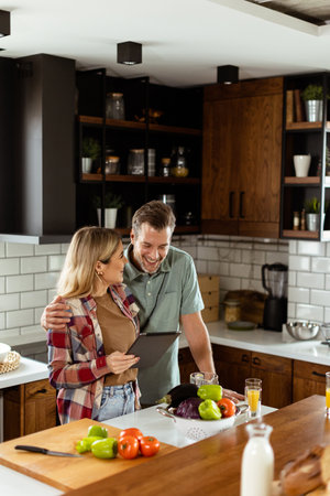 A cheerful couple stands in a well-lit kitchen, engrossed in a digital tablet among fresh ingredientsの写真素材