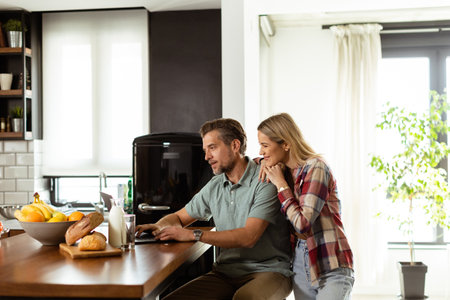 A cheerful couple enjoys a light-hearted moment in their sunny kitchen, working on laptop surrounded by a healthy breakfastの写真素材