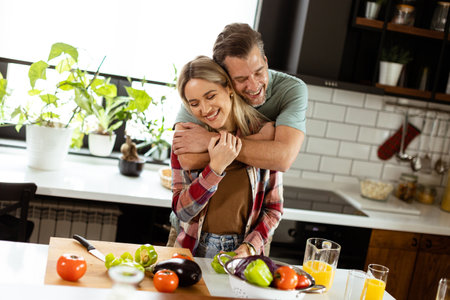 A man and woman share a warm hug in a cozy kitchen, surrounded by cooking utensils and a pot simmering on the stove.の写真素材