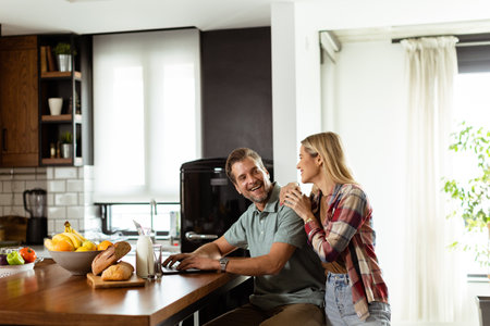 A cheerful couple enjoys a light-hearted moment in their sunny kitchen, working on laptop surrounded by a healthy breakfastの写真素材