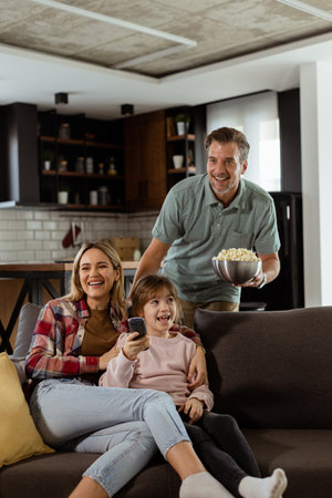 A family of three is comfortably nestled on a couch, their faces reflecting excitement and attentiveness as they share a bowl of popcorn during a suspenseful movie nightの写真素材