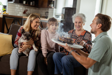 Heartwarming scene unfolds as a multi-generational family gathers on a couch to present a birthday cake to a delighted grandmother, creating memories to cherishの写真素材