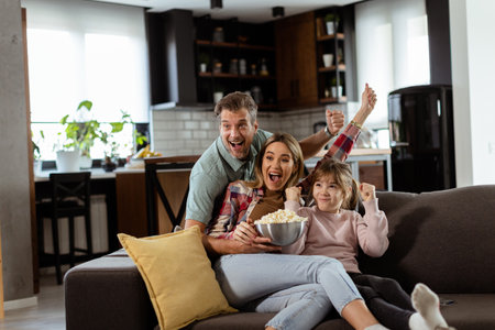 A family of three is comfortably nestled on a couch, their faces reflecting excitement and attentiveness as they share a bowl of popcorn during a suspenseful movie nightの写真素材